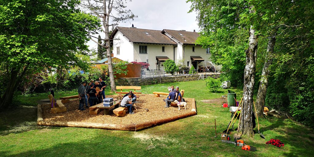 Der Grillplatz wird eingeweiht. Menschen sitzen auf den Holzbänken und stehen um den Grill.